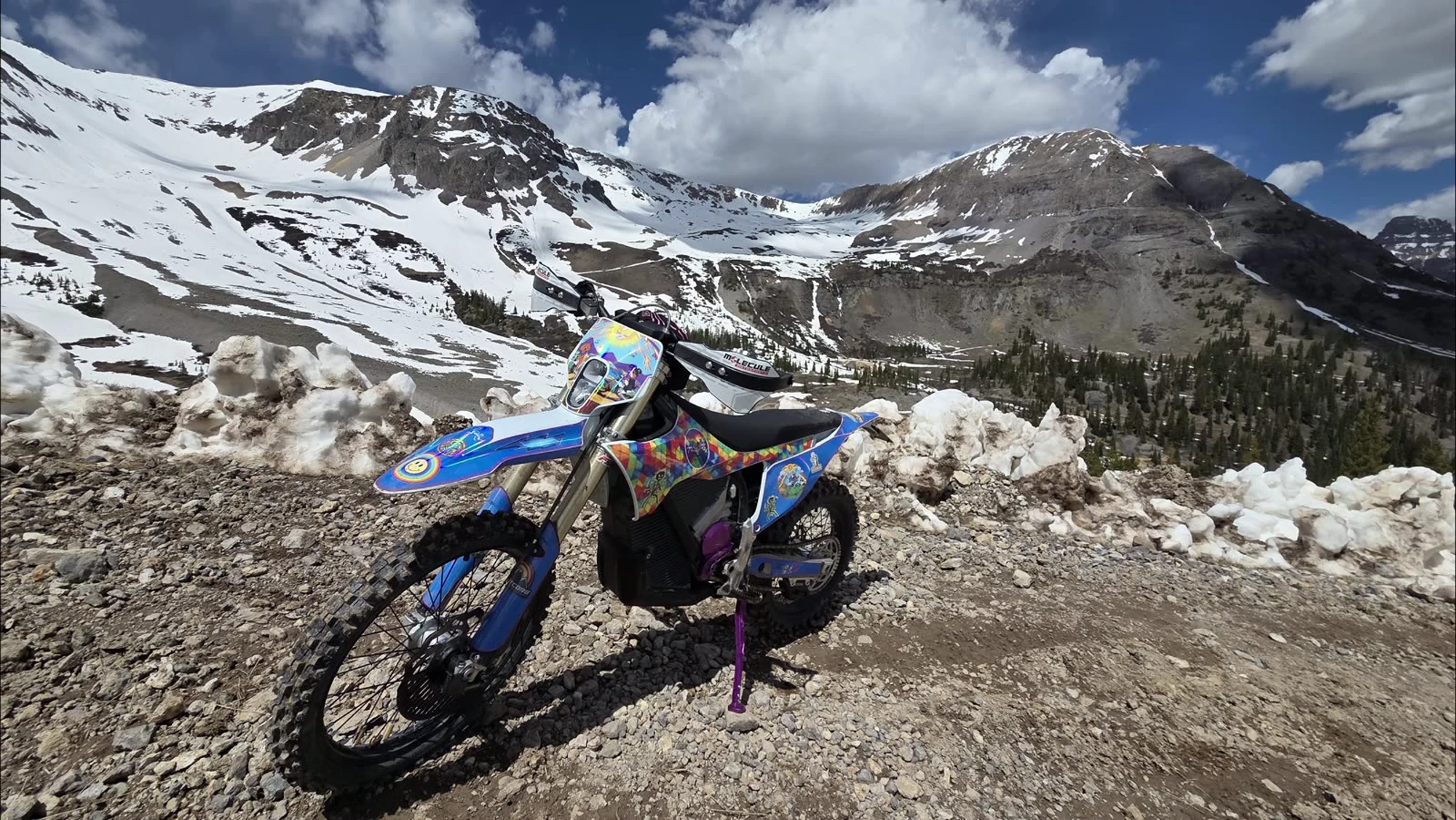 An electric dirt bike parked on a rocky pass, snow-covered San Juan peaks behind it.