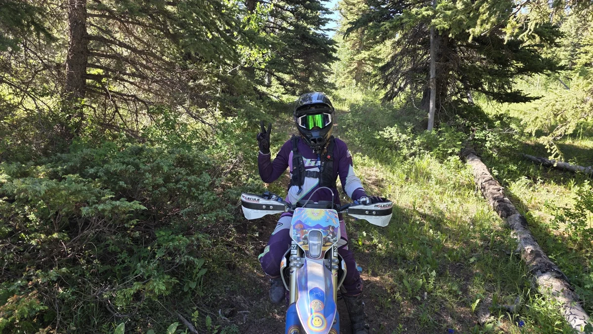 Colleen on her bike in the trees, purple gear, throwing a peace sign.