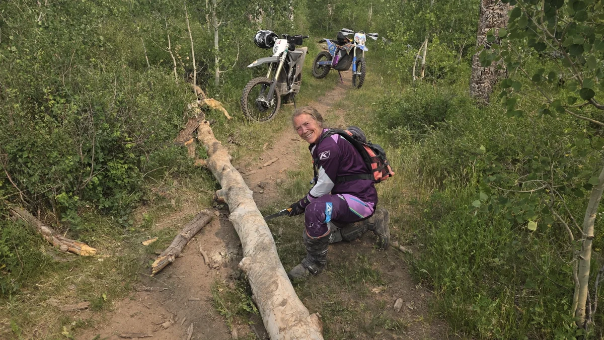 Colleen kneeling on a trail with a hand saw, cutting through a fallen aspen. Two bikes parked behind her.
