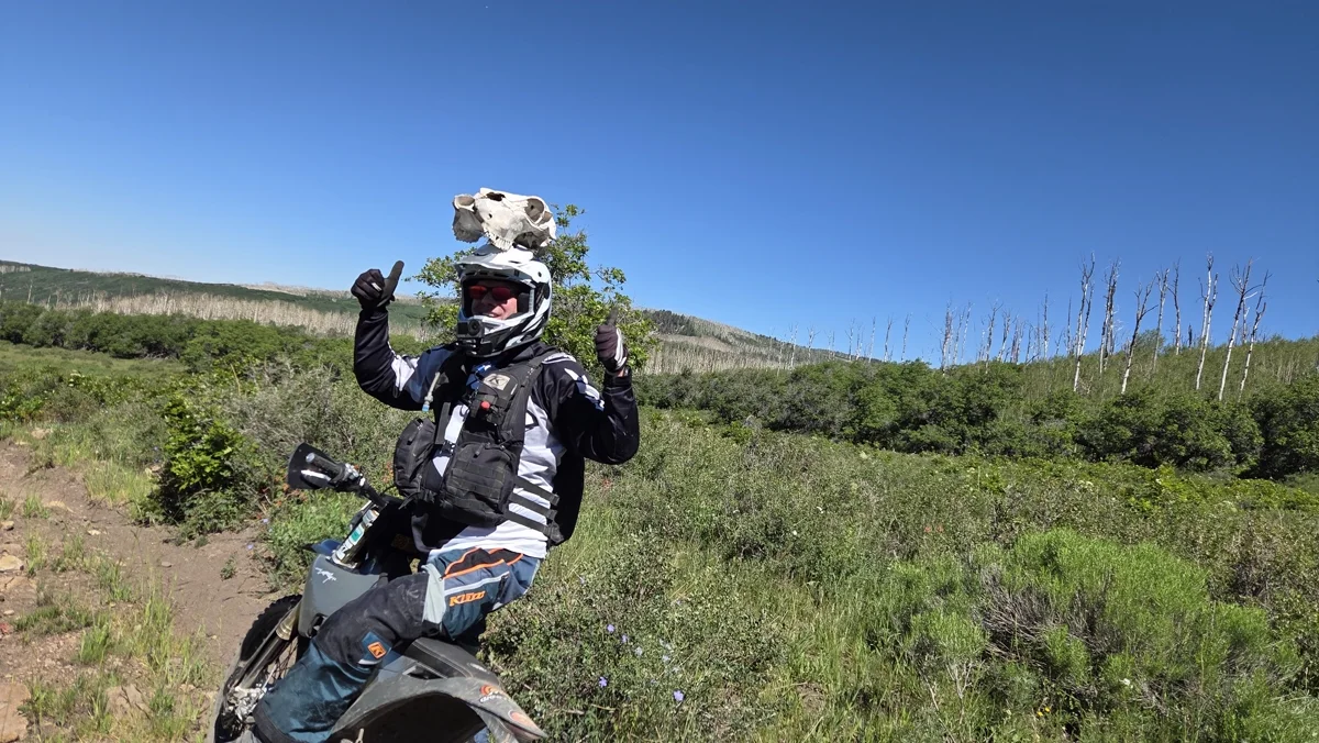 A rider on a dirt bike with an animal skull balanced on top of his helmet, giving two thumbs up.
